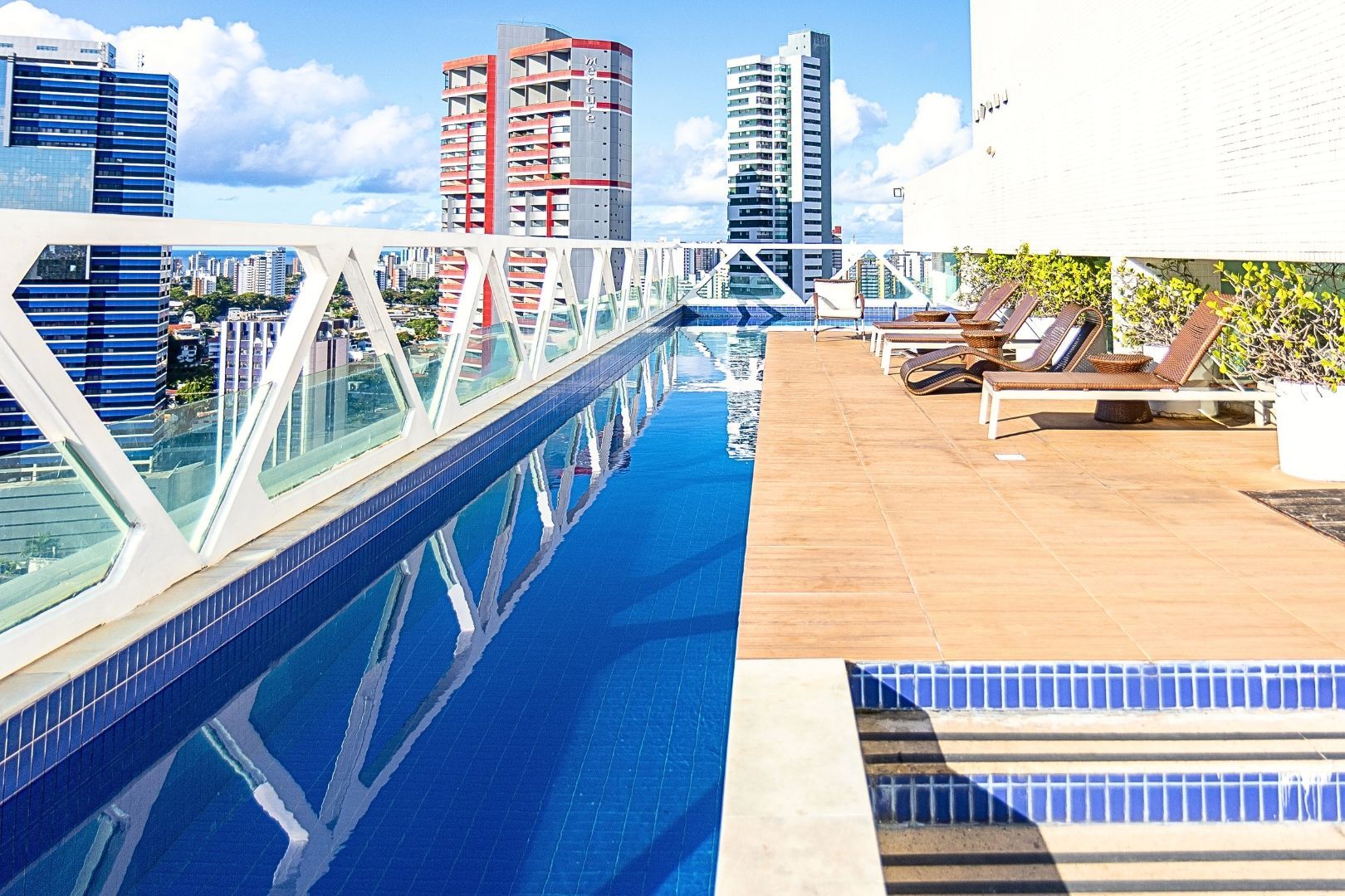 Piscina de borda infinita no terraço, com água azul, guarda-corpo de vidro, deck de madeira, espreguiçadeiras e prédios da cidade ao fundo
