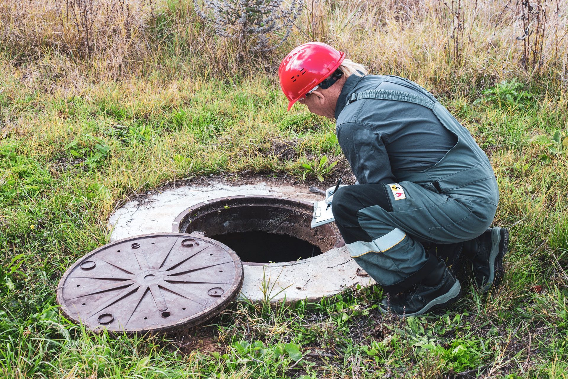 Worker in red helmet inspecting an open manhole cover in a grassy field