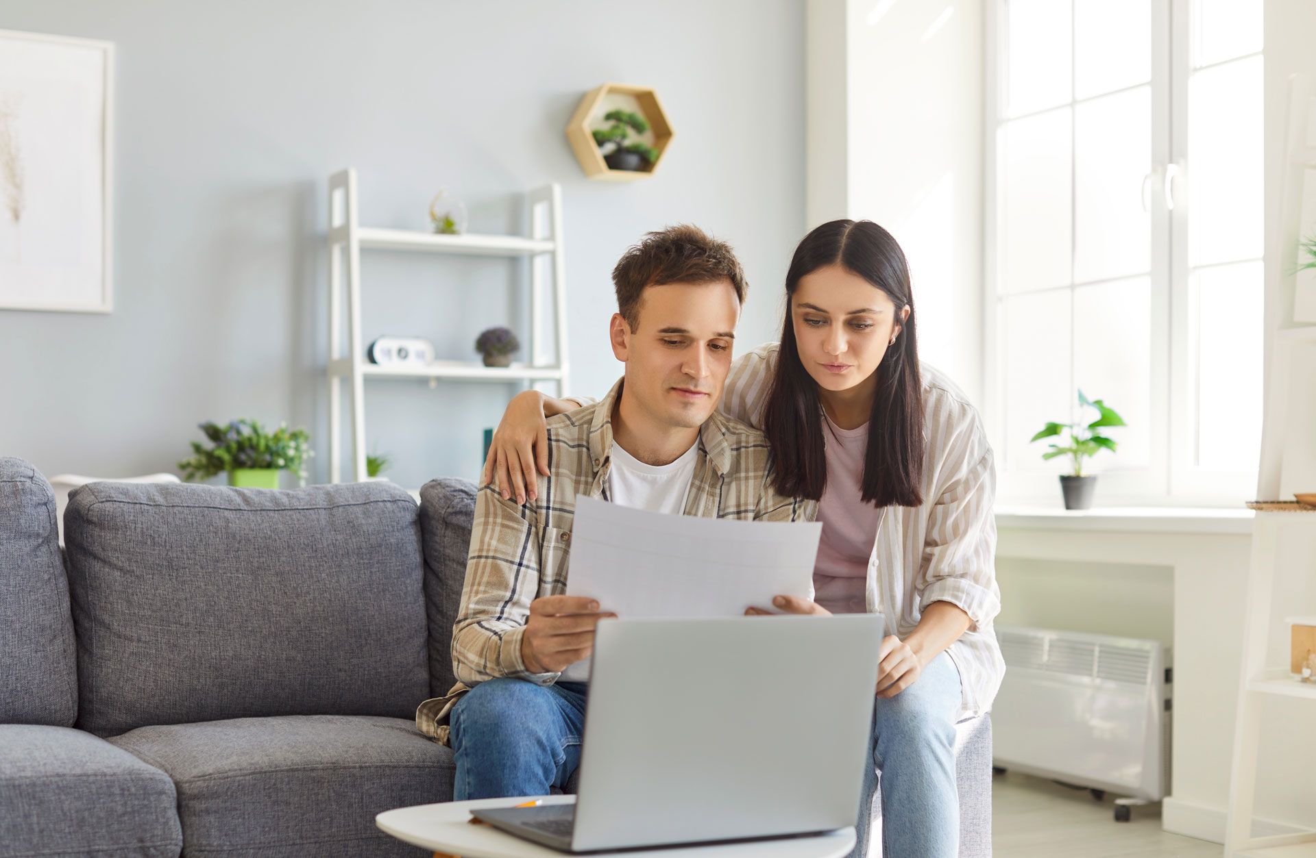 Two people reviewing a document beside a laptop on a couch in a bright living room