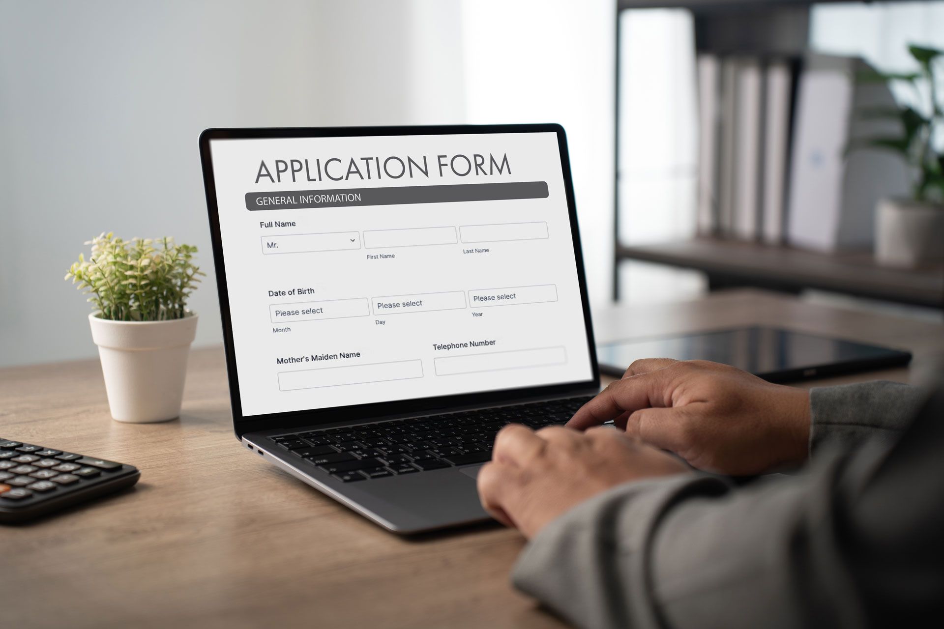 Person filling out an application form on a laptop at a desk with a plant and calculator nearby