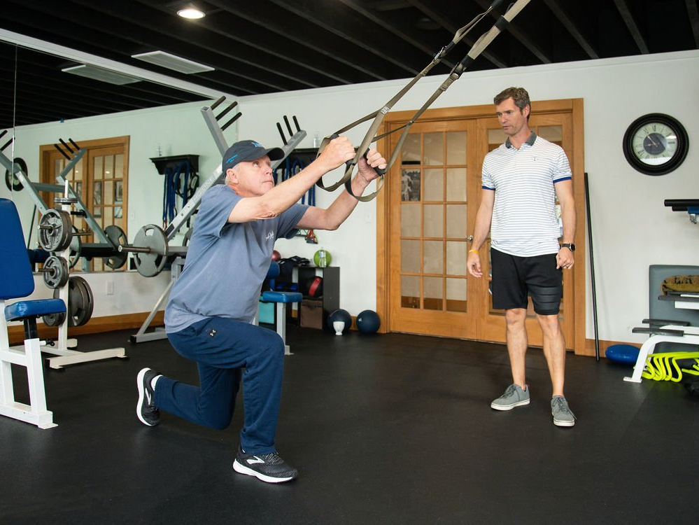 A man is doing trx exercises in a gym while another man watches.