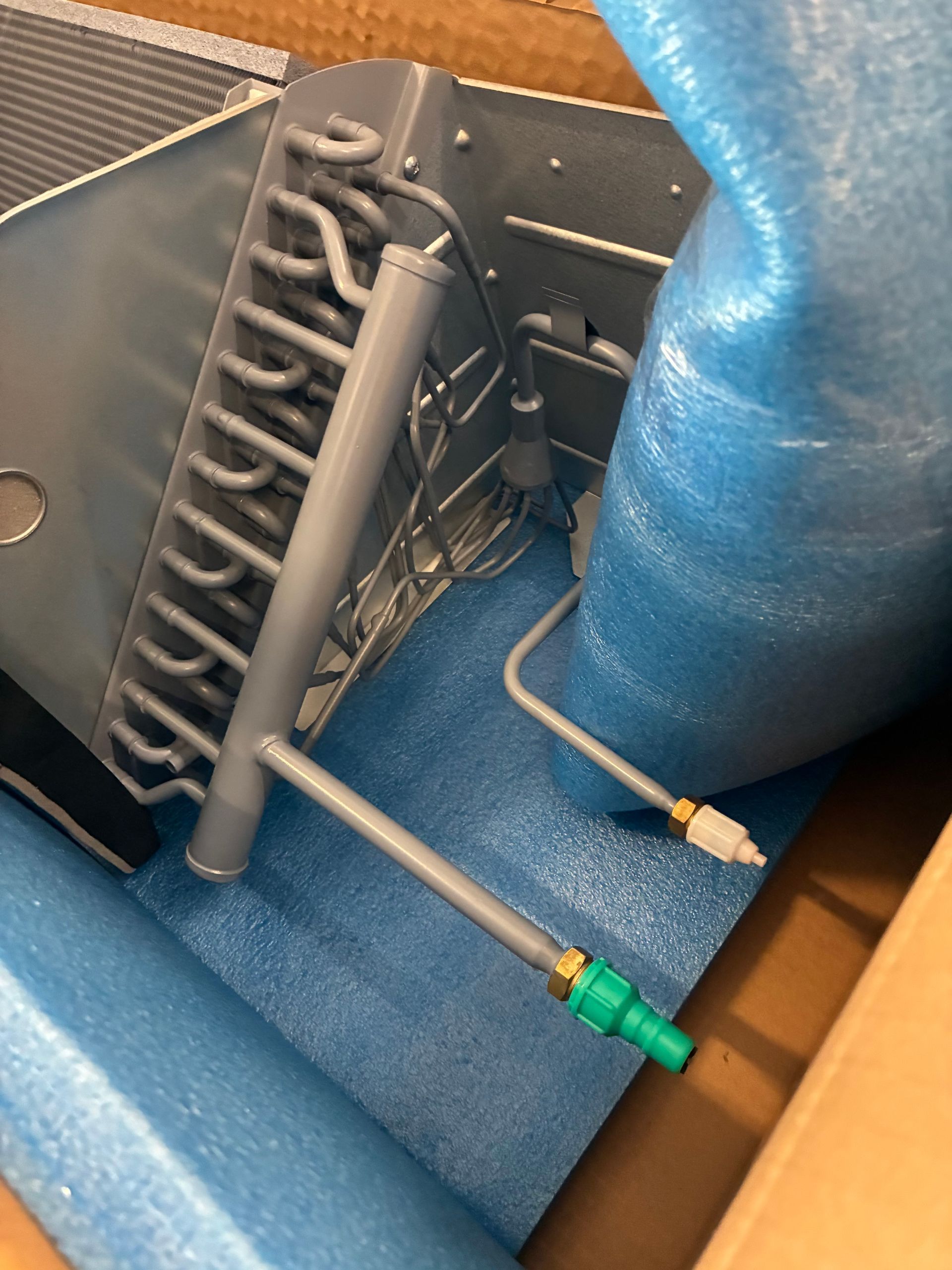 A person in a blue glove uses a spray nozzle to clean a blue industrial heat exchanger coil in a factory.