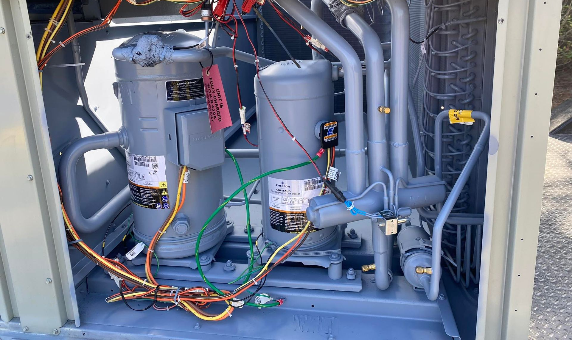 An open HVAC unit cabinet showing two grey vertical compressors connected by pipes and wiring against a metal frame.