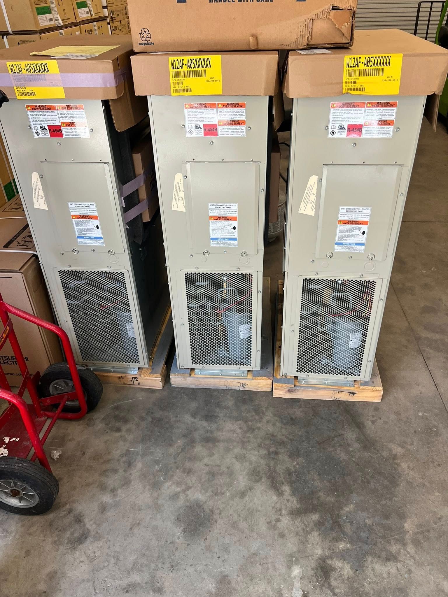 Three tall, beige industrial cooling units stand on pallets in a warehouse, with boxes stacked nearby and a hand truck.