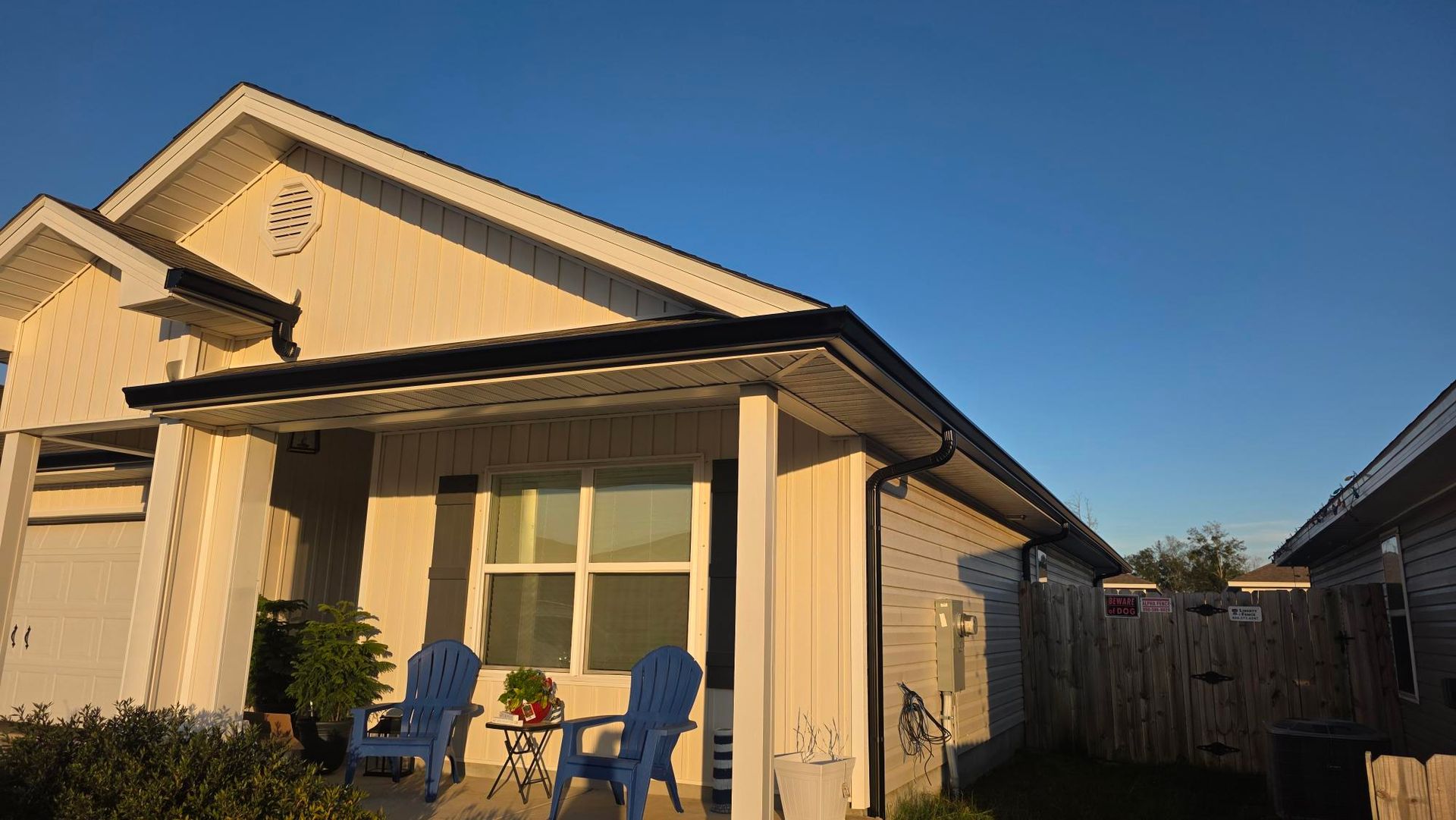 A cream-colored house with a dark roof and a small porch. Two blue chairs sit outside the window.