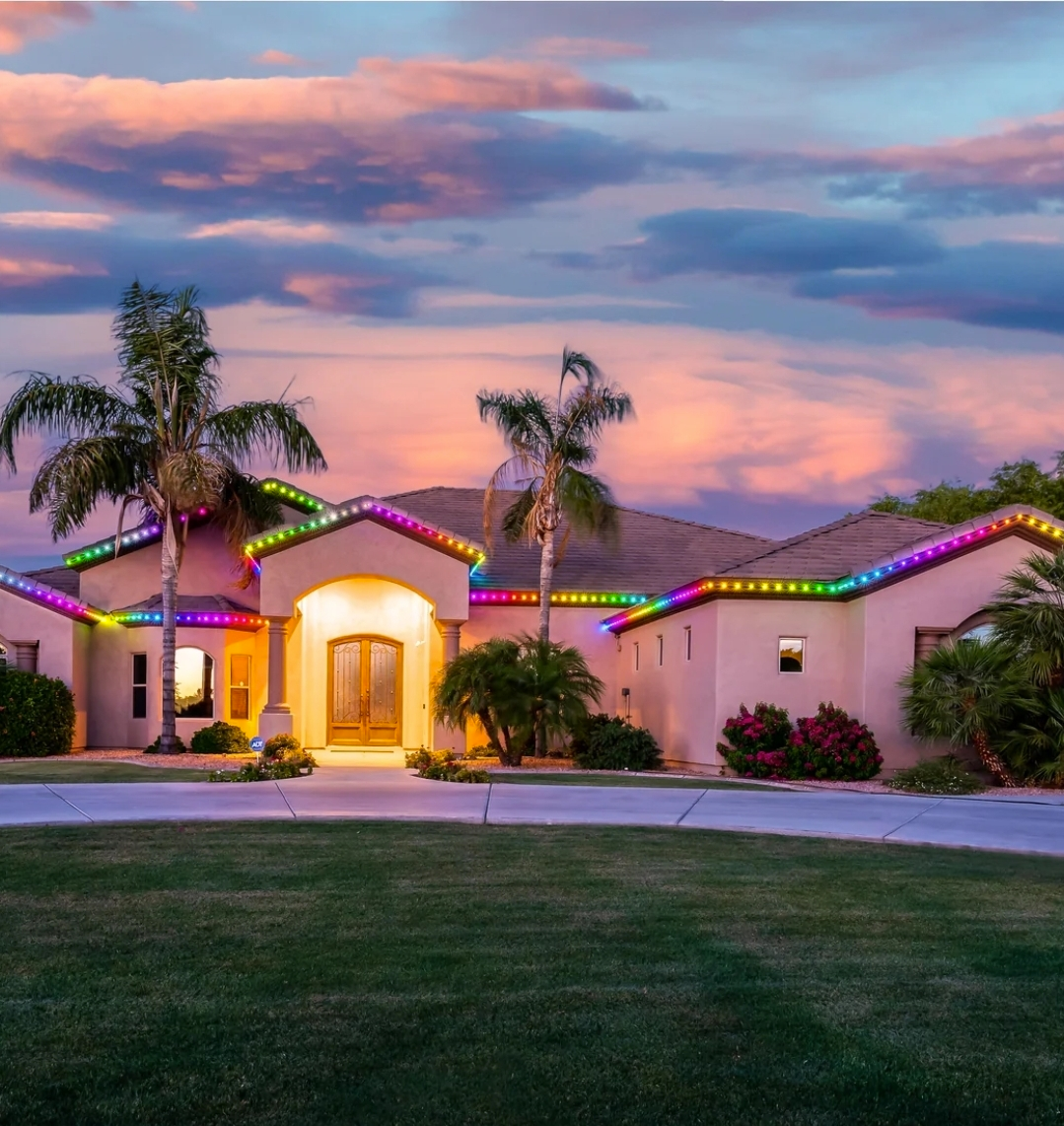 House with rainbow-colored lights outlining the roof, under a colorful sunset sky. Palm trees in front.