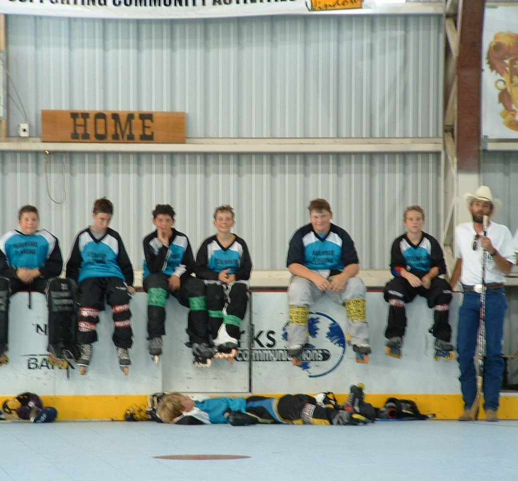 A row of roller hockey players in teal and black jerseys sit on the rink boards next to a coach in a cowboy hat.