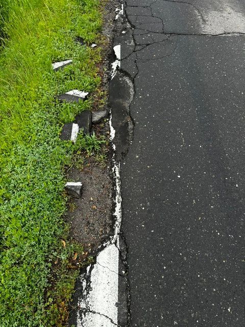 A section of a cracked, deteriorating asphalt road edge with broken white lane markings alongside a grassy roadside.
