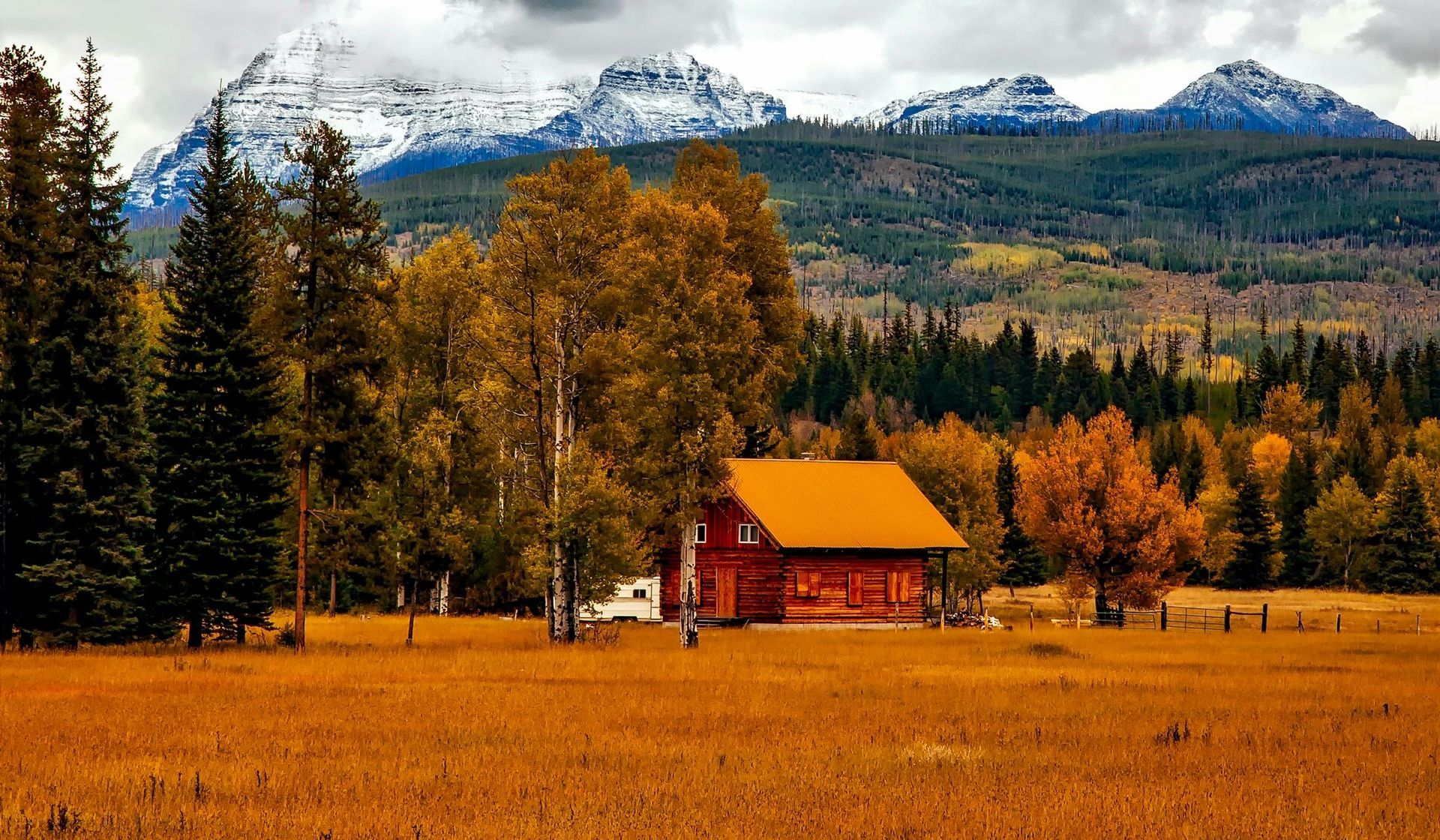 Log cabin in a golden field with autumn trees and snowy mountains in the background.