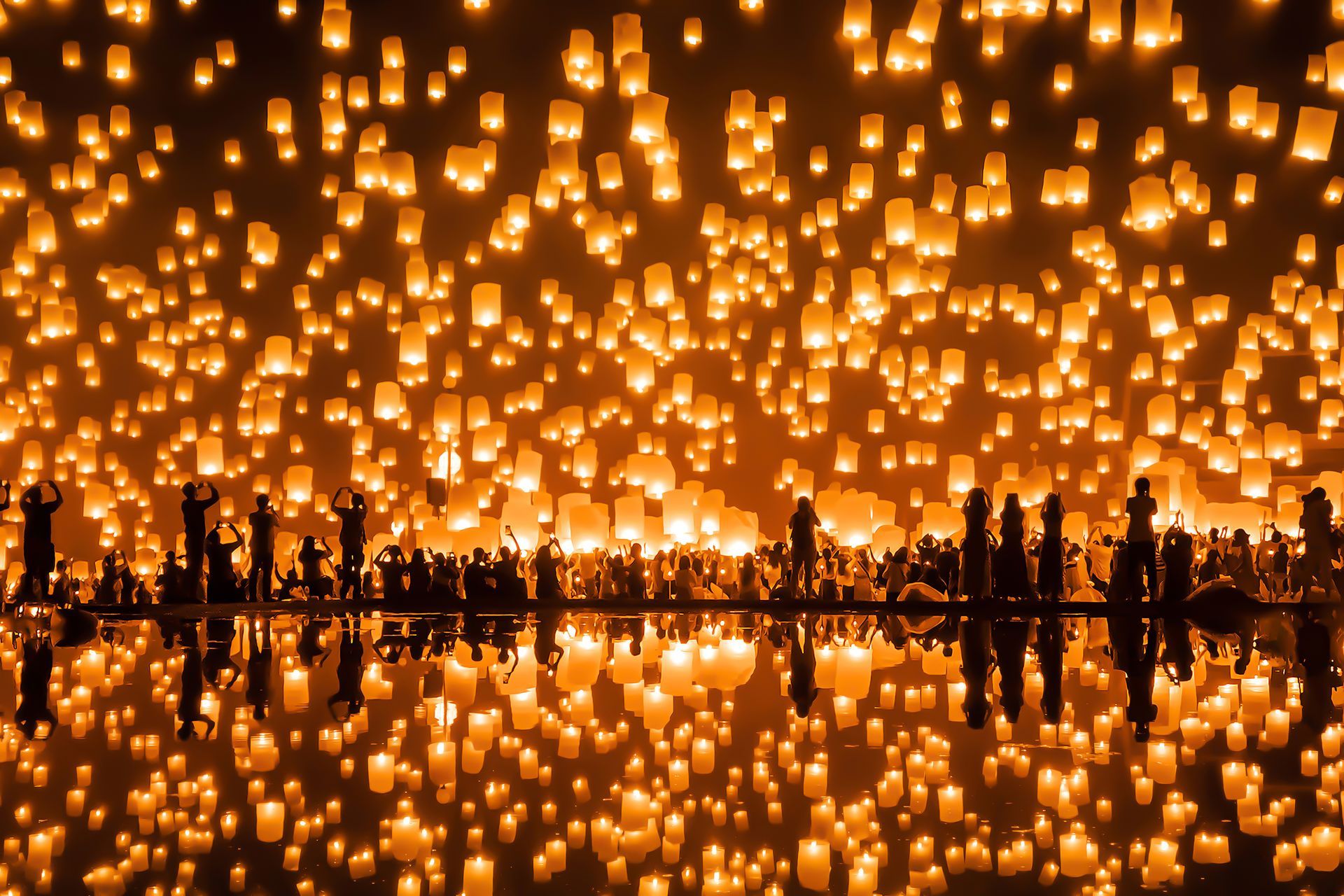 Silhouettes of people amid glowing lanterns, reflected in calm water at night