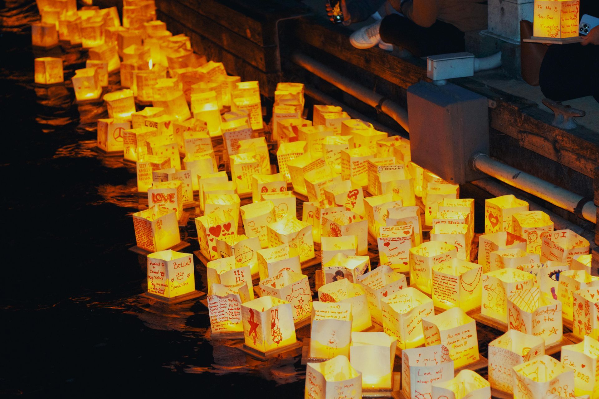 Rows of glowing lanterns lit in warm orange light along a dark walkway or canal edge
