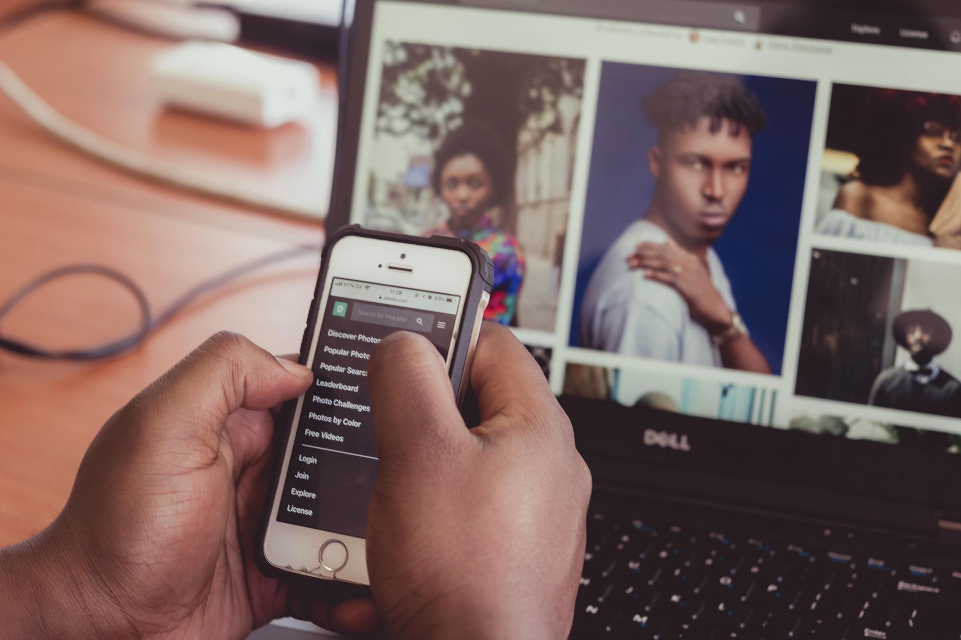 Hands holding a phone in front of a Dell laptop displaying portrait photos and a search list