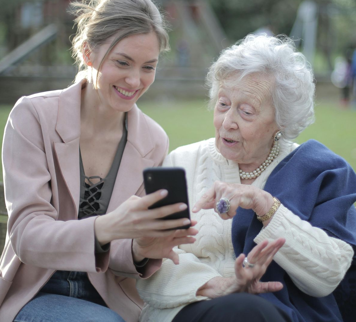 Two women sitting together outdoors, smiling at a smartphone and gesturing at the screen