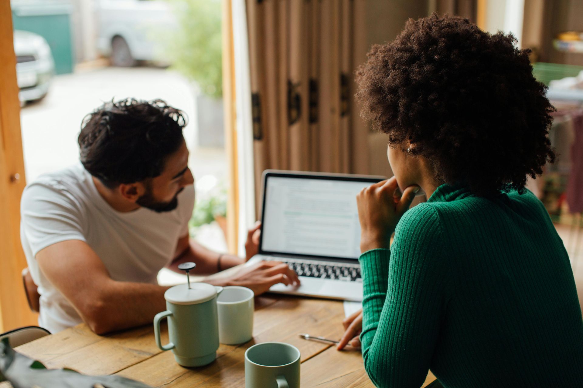 Two people collaborating at a table, looking at a laptop in a bright cafe-like setting.