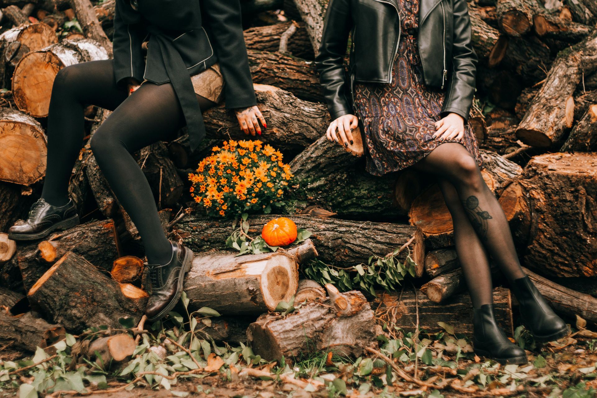 Two people sit on stacked logs beside orange flowers and a small pumpkin in a wooded setting