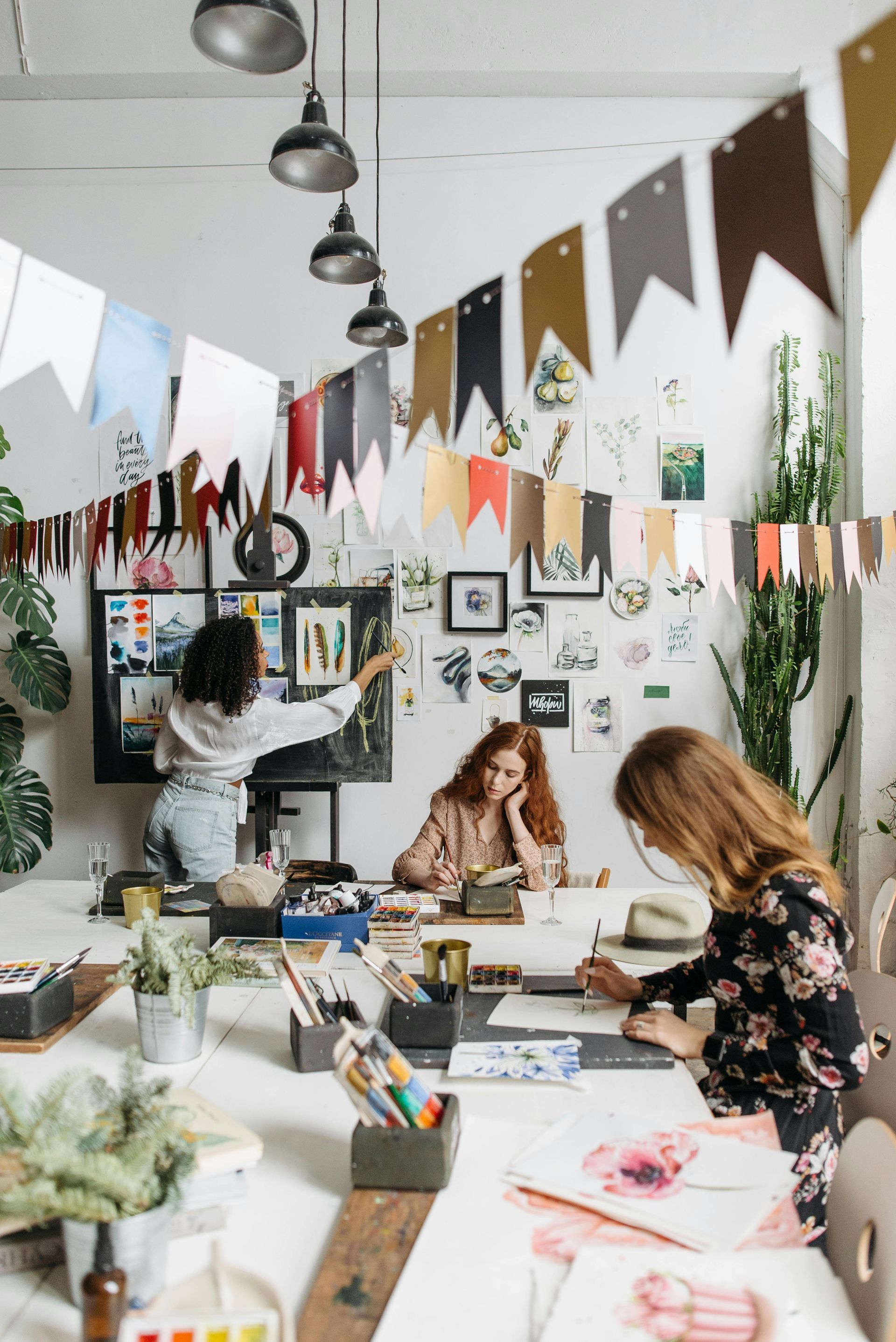 Art studio classroom with people painting at tables, colorful bunting, and framed artwork on the walls
