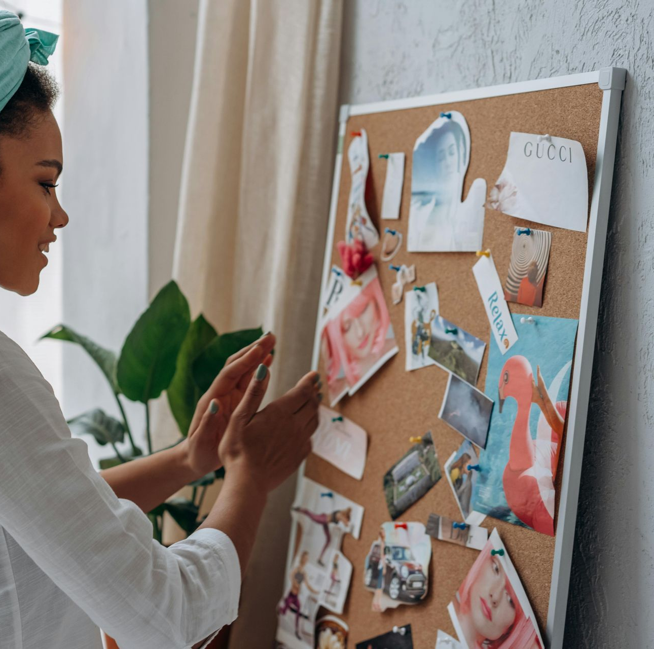 Person pinning photos and notes on a corkboard beside a plant in a bright room