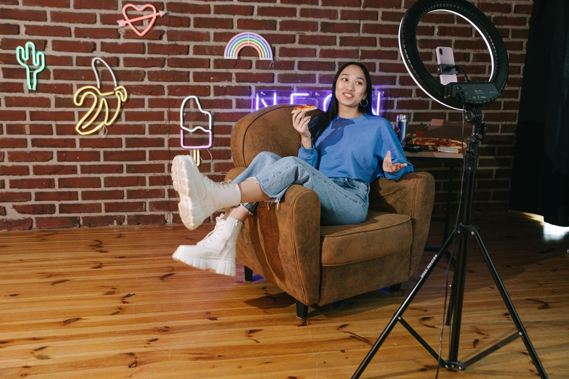 Woman sitting on a brown armchair in a studio, with a ring light and neon wall decor on a brick wall background