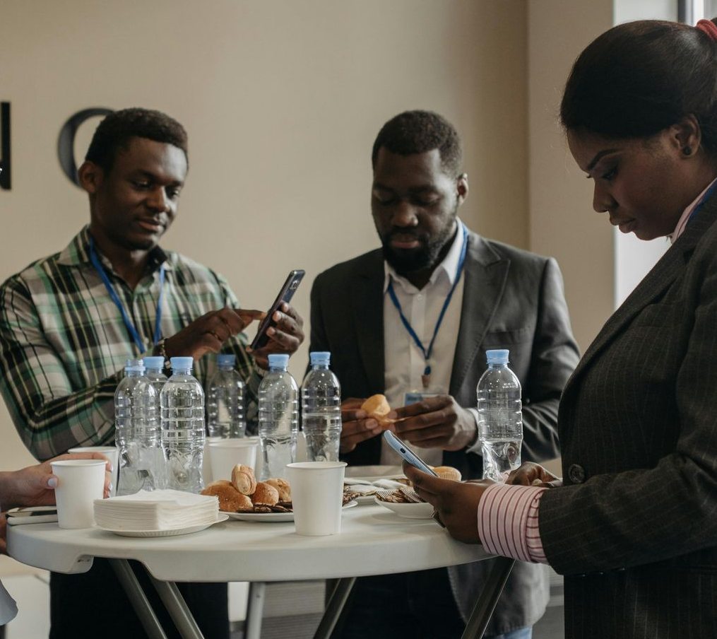 Three people standing at a table with water bottles, cups, and pastries, looking at phones and papers
