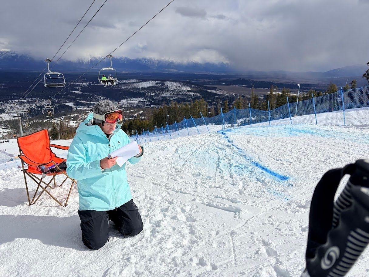 Person kneels on snowy ski slope reading a map, with mountain view, chairlifts, and orange chair nearby