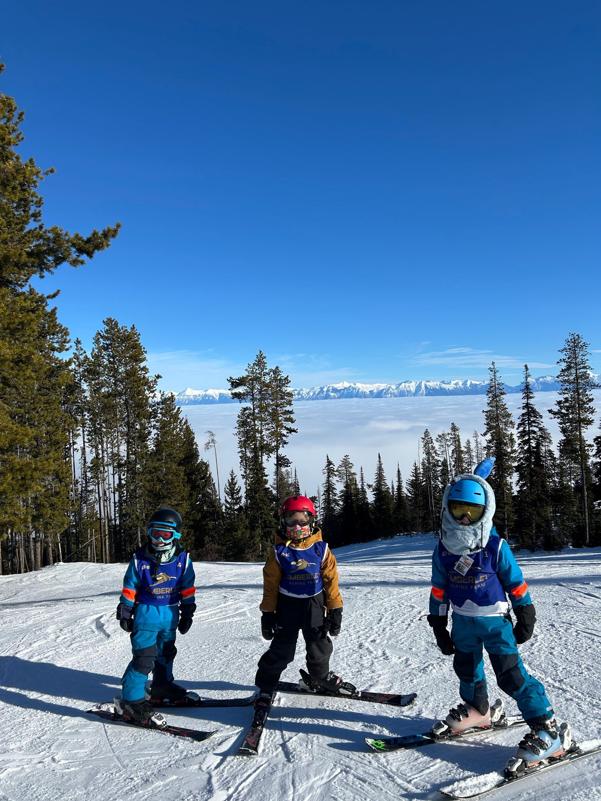 Kimberley Alpine Team Three skiers in colorful jackets on a snowy mountain trail under a blue sky.