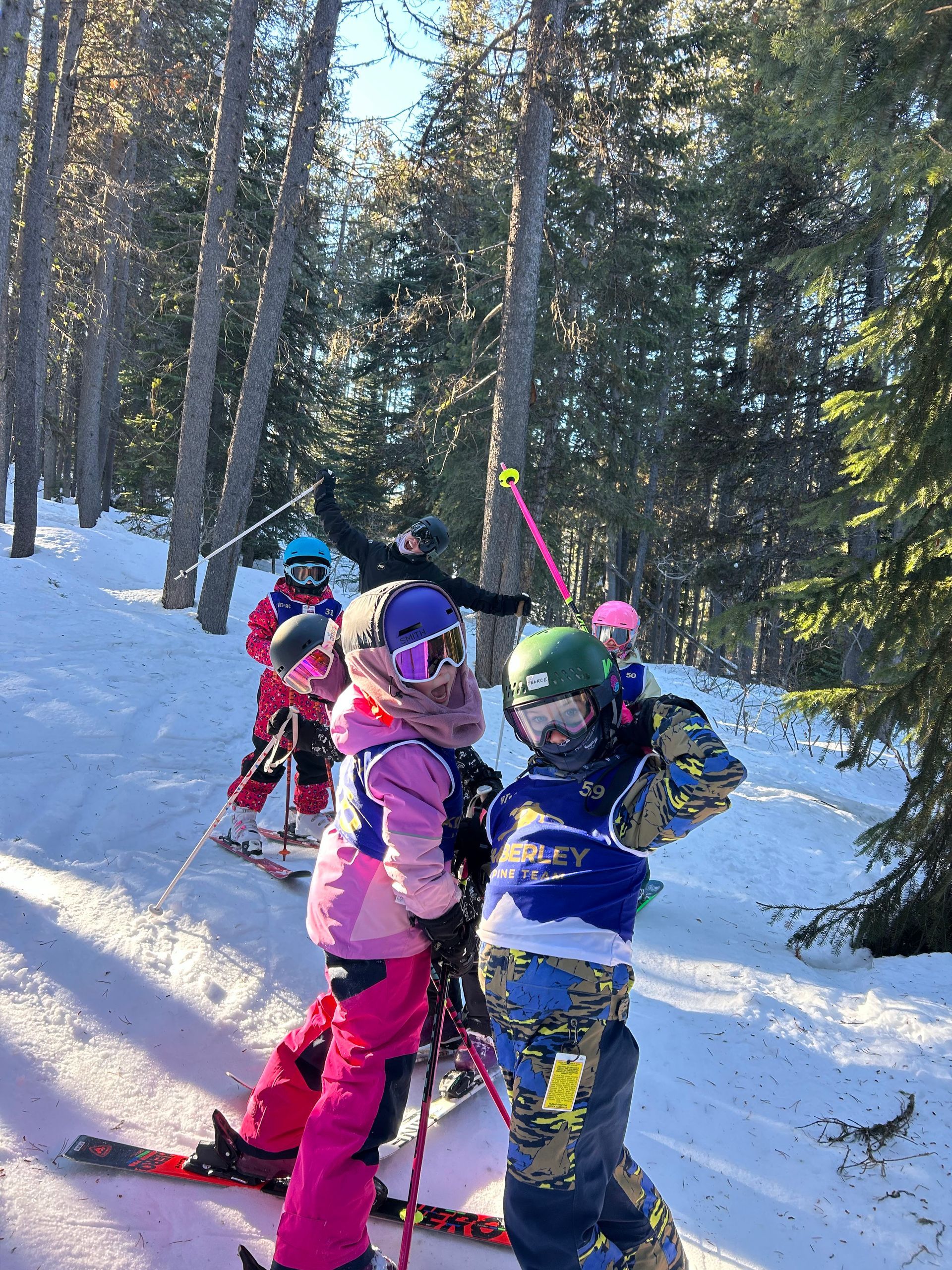 Kimberley Alpine Team Children in colorful ski gear stand on a snowy trail in a forest, holding ski poles.