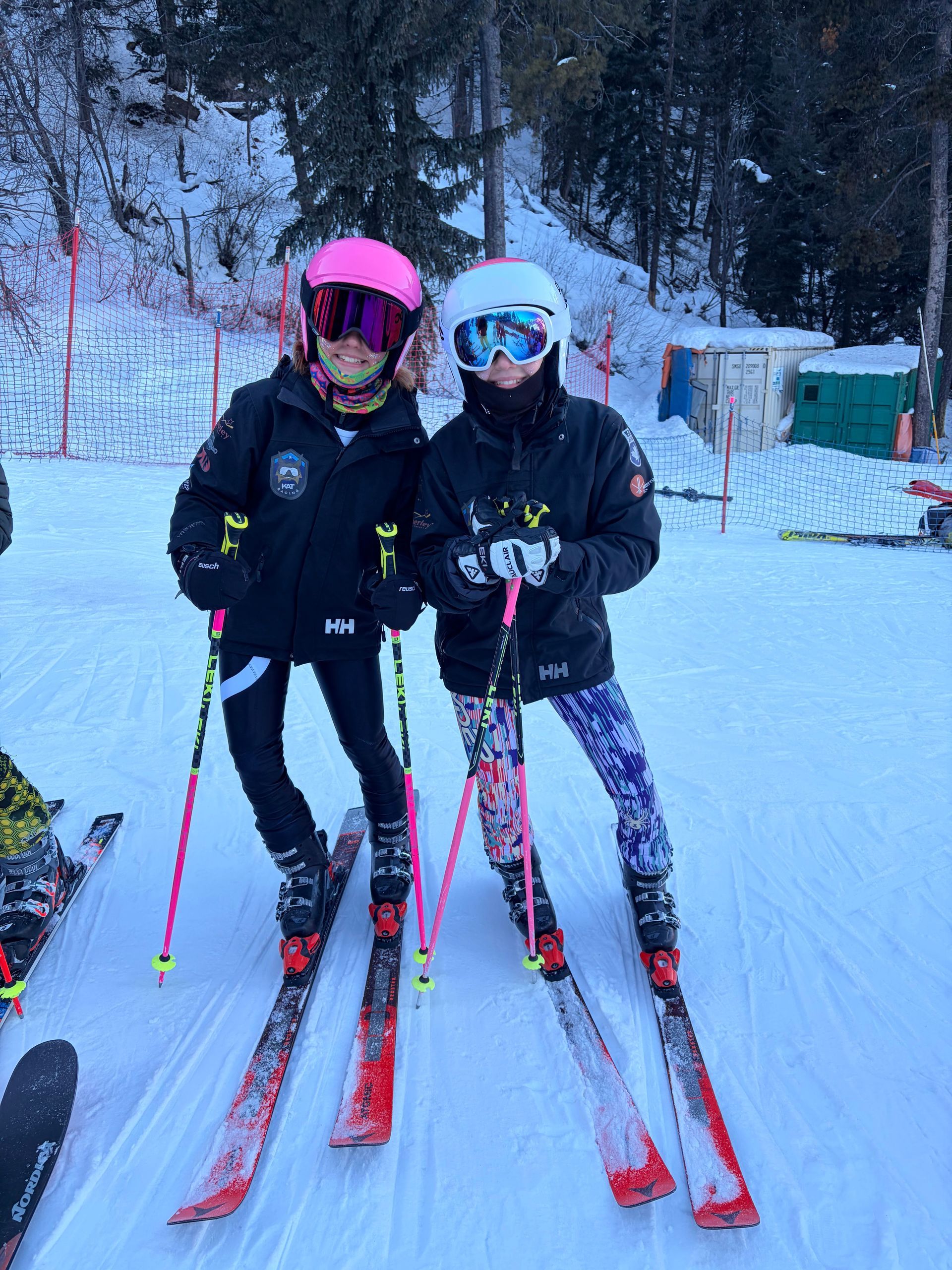 Kimberley Alpine Team Two skiers in helmets and goggles pose with skis on a snowy trail in a winter forest.