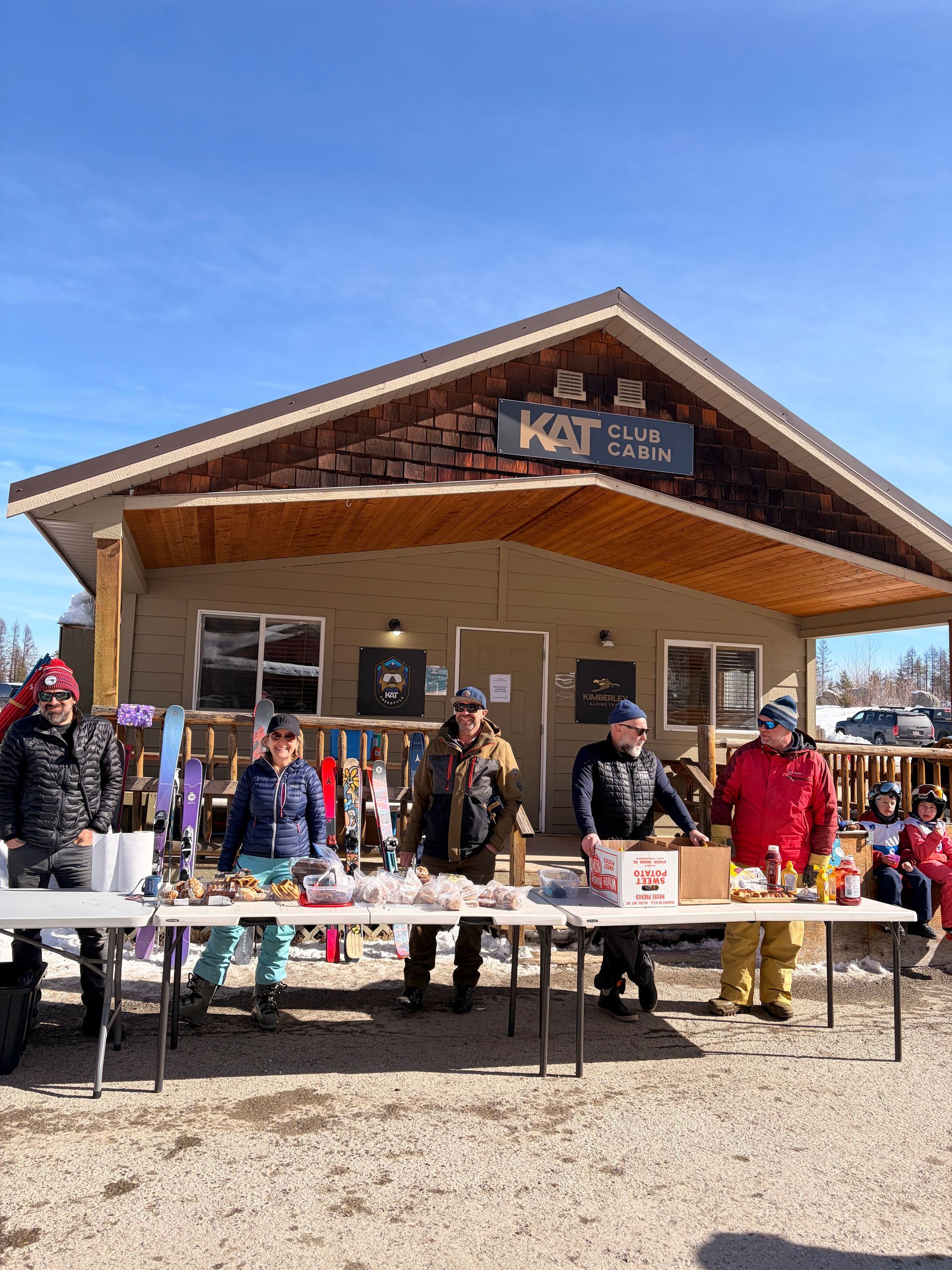 Kimberley Alpine Team People standing behind tables outside a small wooden building under a blue sky
