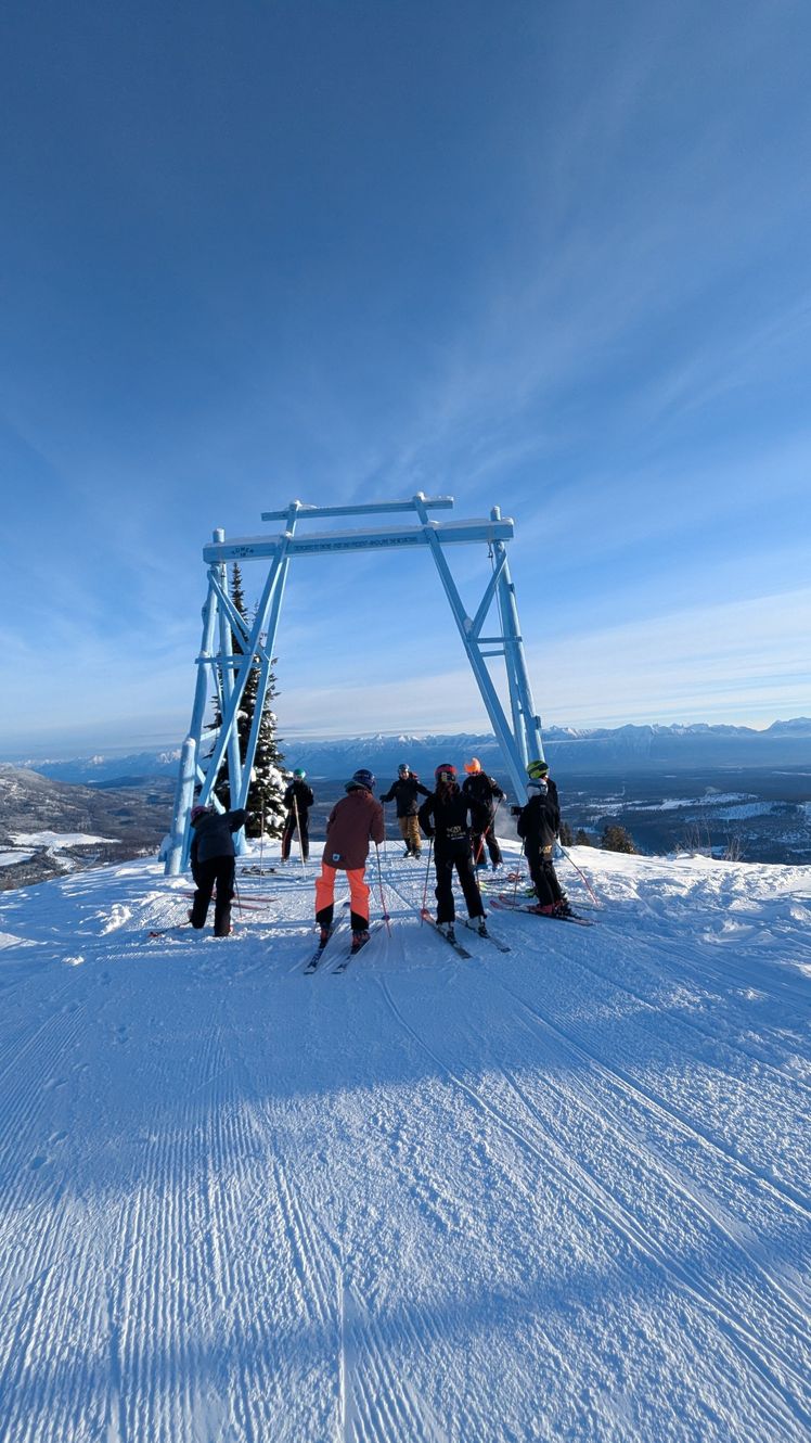 Kimberley Alpine Team People standing on a snowy mountain summit beside a blue ski-lift structure under a clear sky