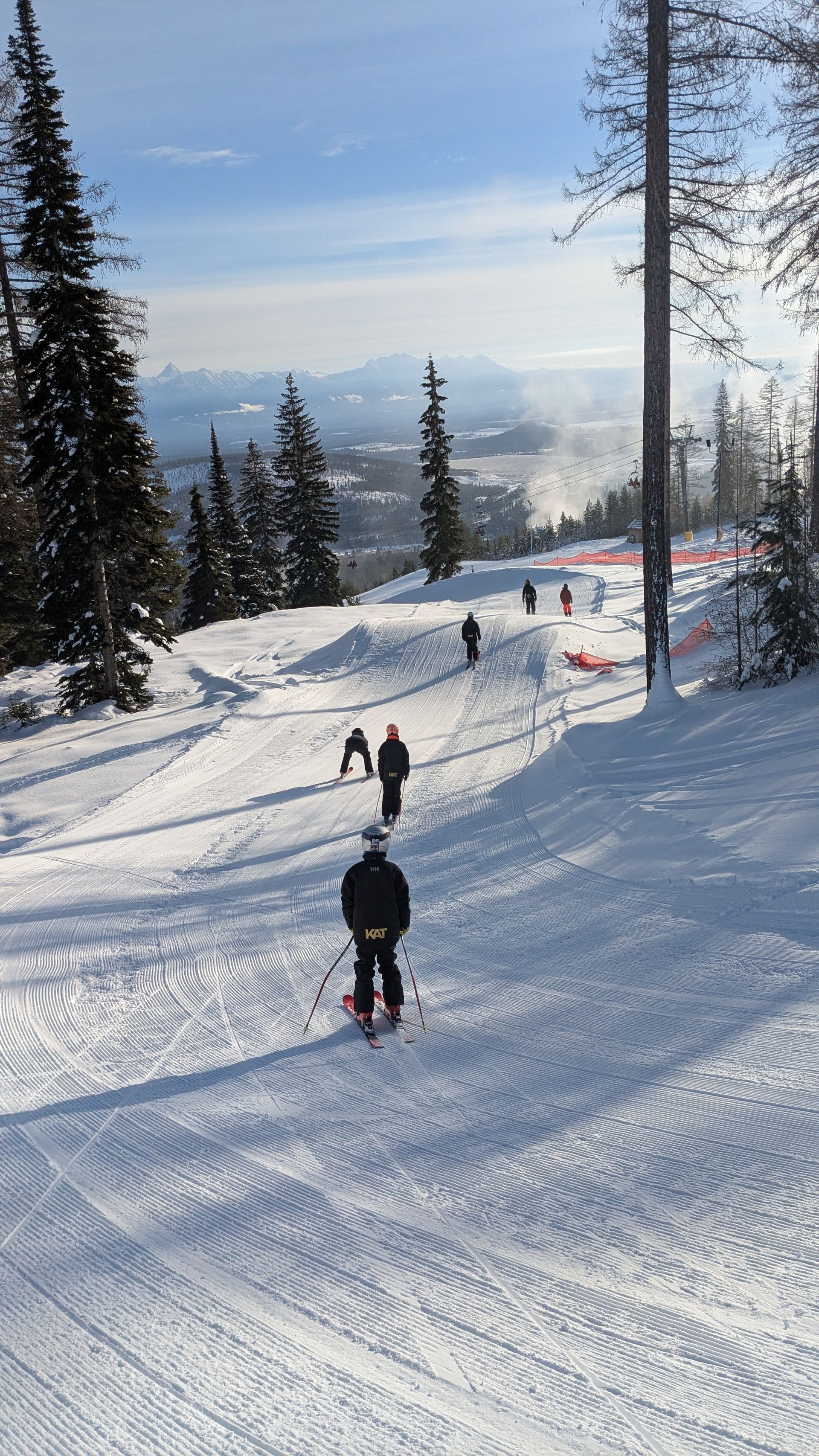 Kimberley Alpine Team Skiers descending a snowy slope under a clear sky, with trees and a valley in the distance