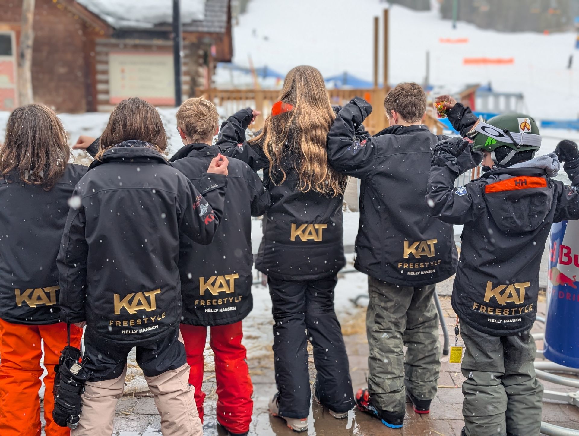 Kimberley Alpine Team Children in black jackets and colorful pants standing outdoors, facing away in a line.