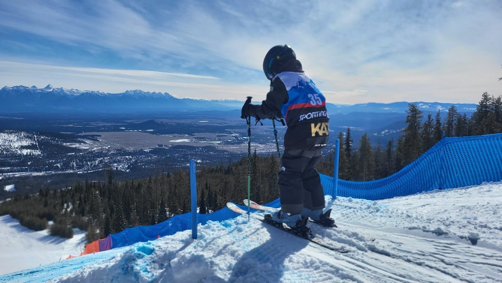 Kimberley Alpine Team Skier in black jacket overlooking a snowy mountain valley under a blue sky
