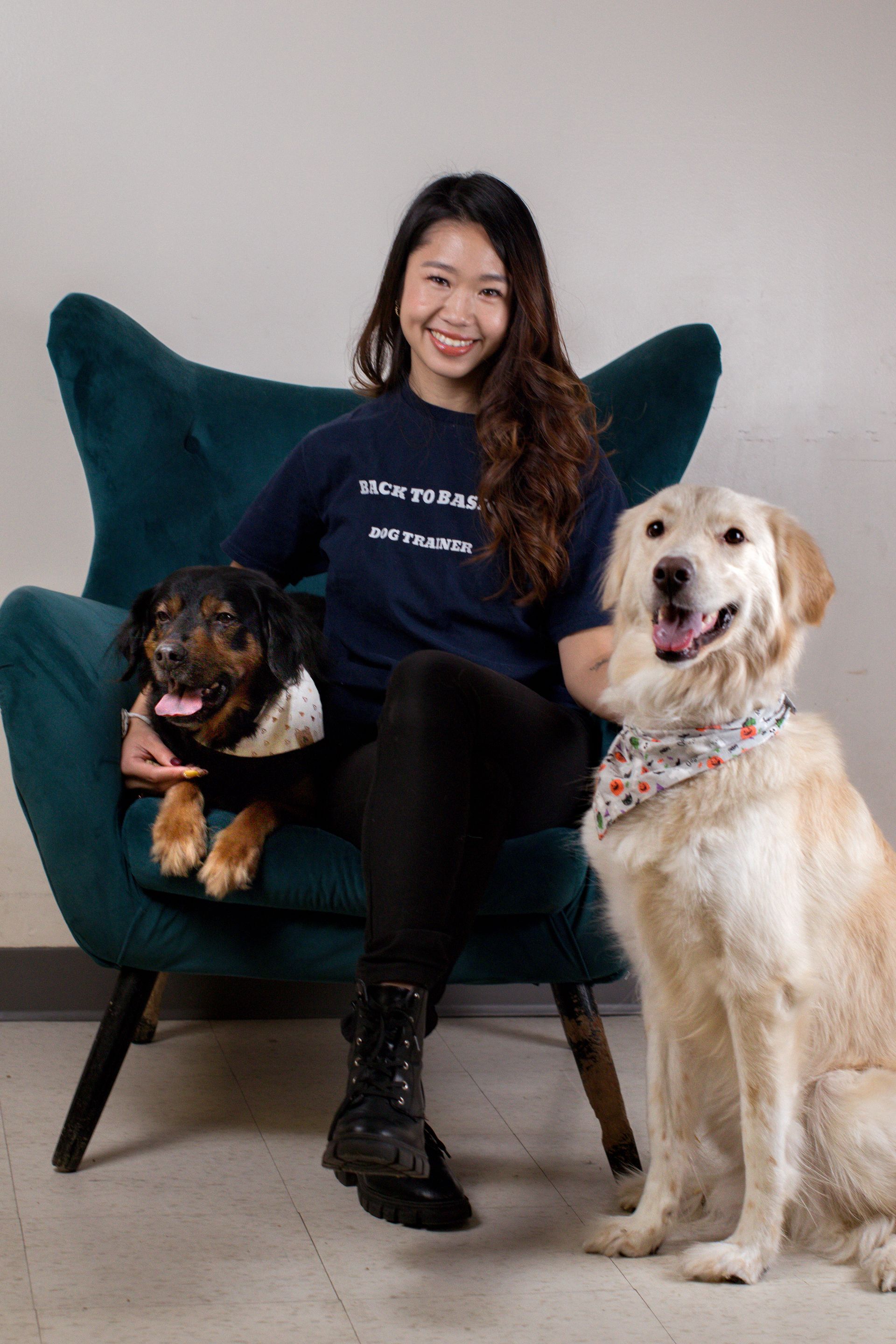 Woman seated in a teal chair with two dogs: a black and brown dog in her lap and a golden retriever beside her.