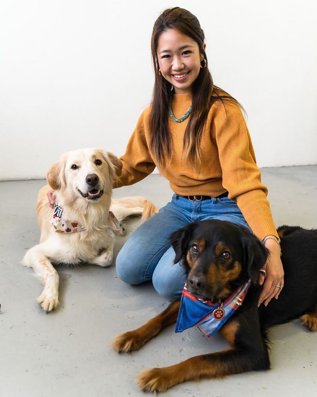 Woman kneeling with two dogs. The dogs wear bandanas. All three are smiling in a white room.