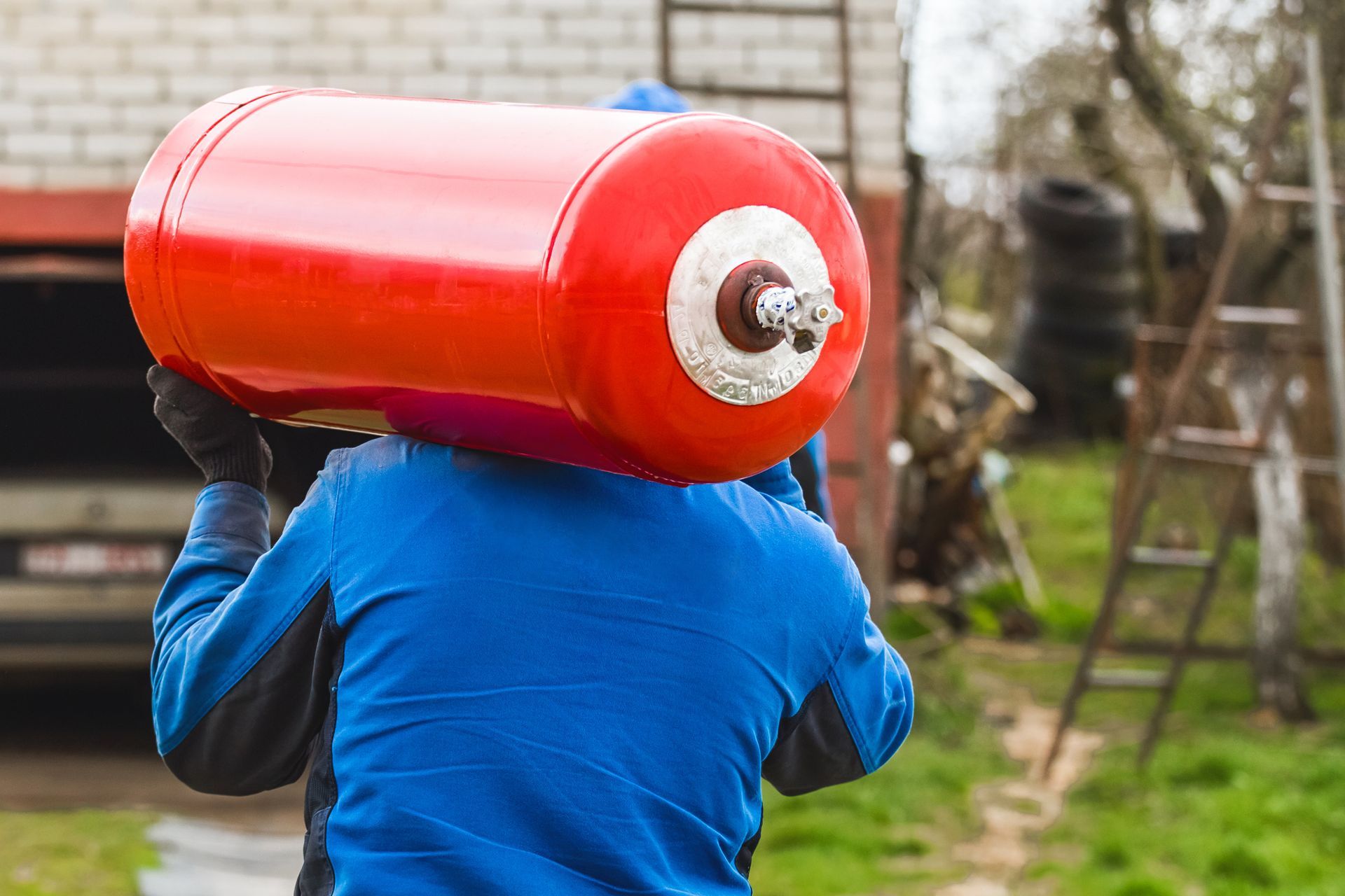 A person wearing a blue jacket carrying a large red gas cylinder on their shoulder outdoors.