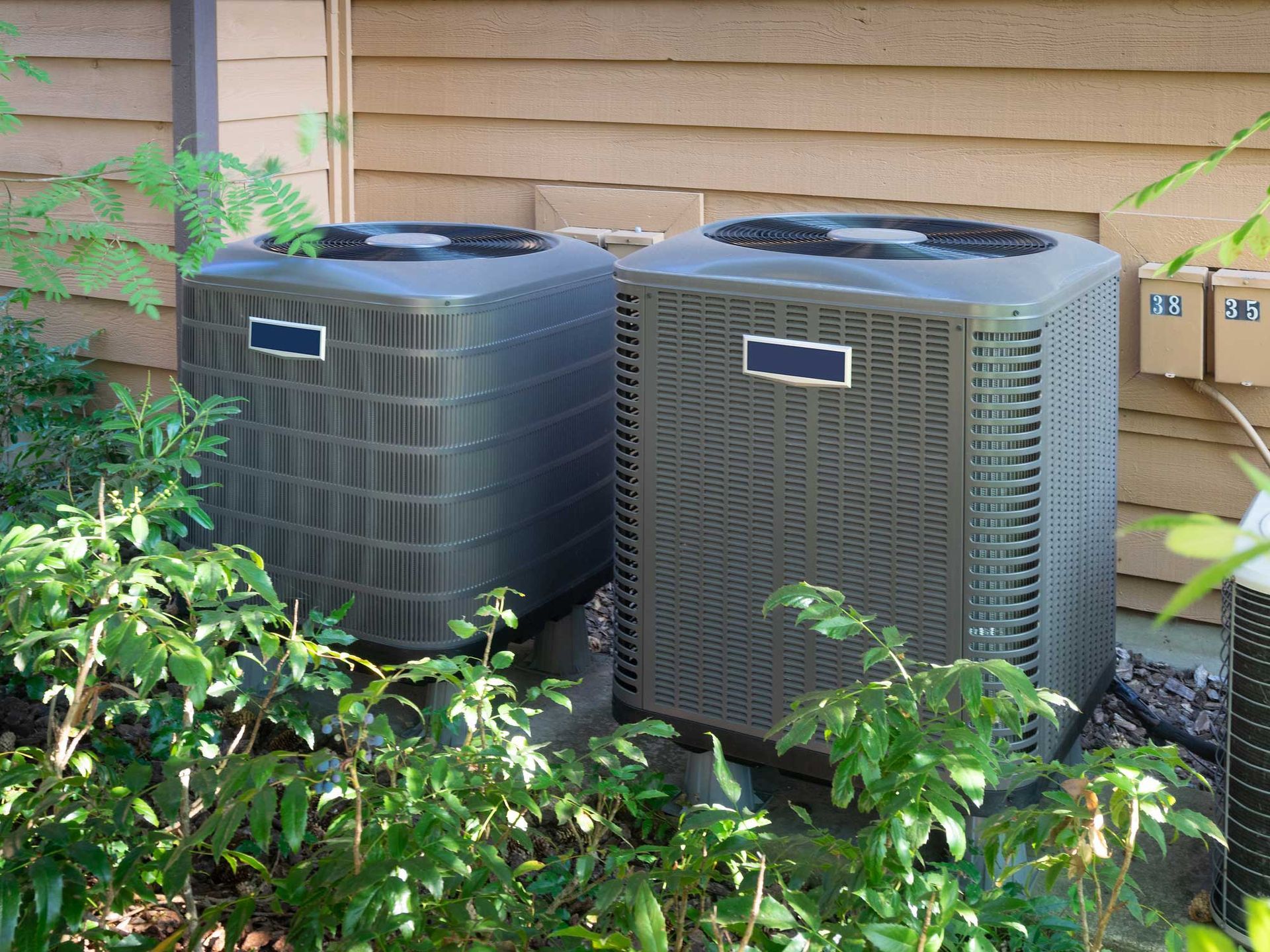 Two gray air conditioning units near bushes against a wood-paneled wall.