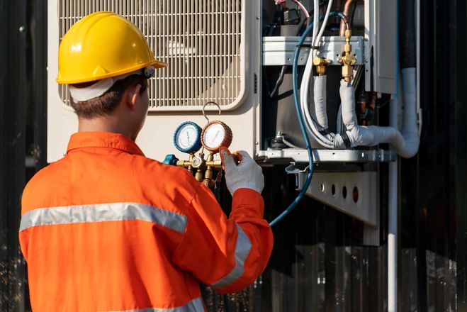 HVAC technician in orange jumpsuit and hard hat checks gauges on an outdoor unit.