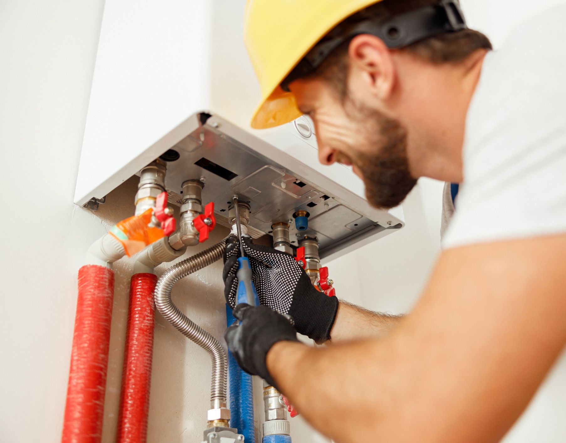 A person in a hard hat working on pipes connected to a white water heater on a wall.