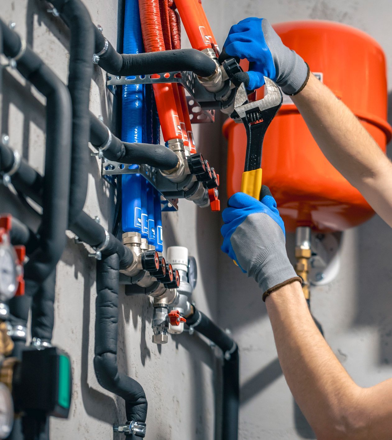 Plumber in blue gloves uses wrench on plumbing pipes with orange tank in background.