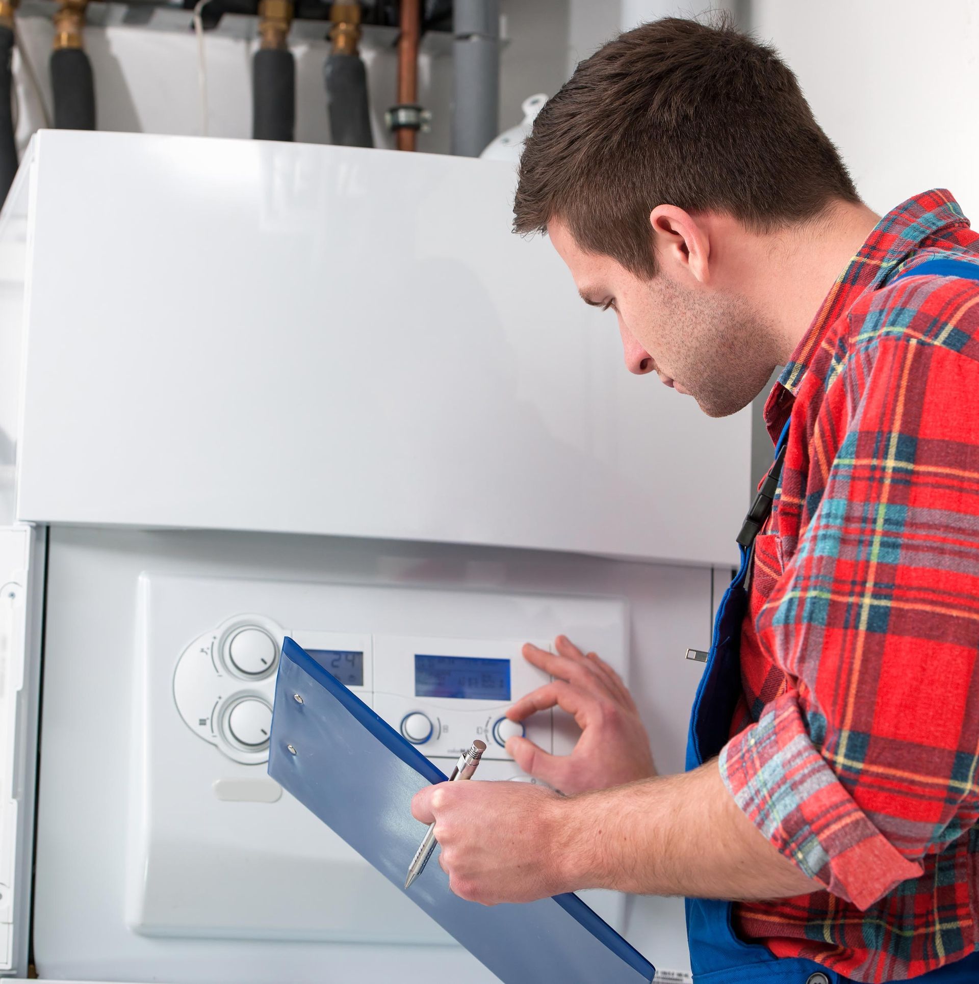 A man in a plaid shirt and overalls inspects a white boiler, holding a clipboard.