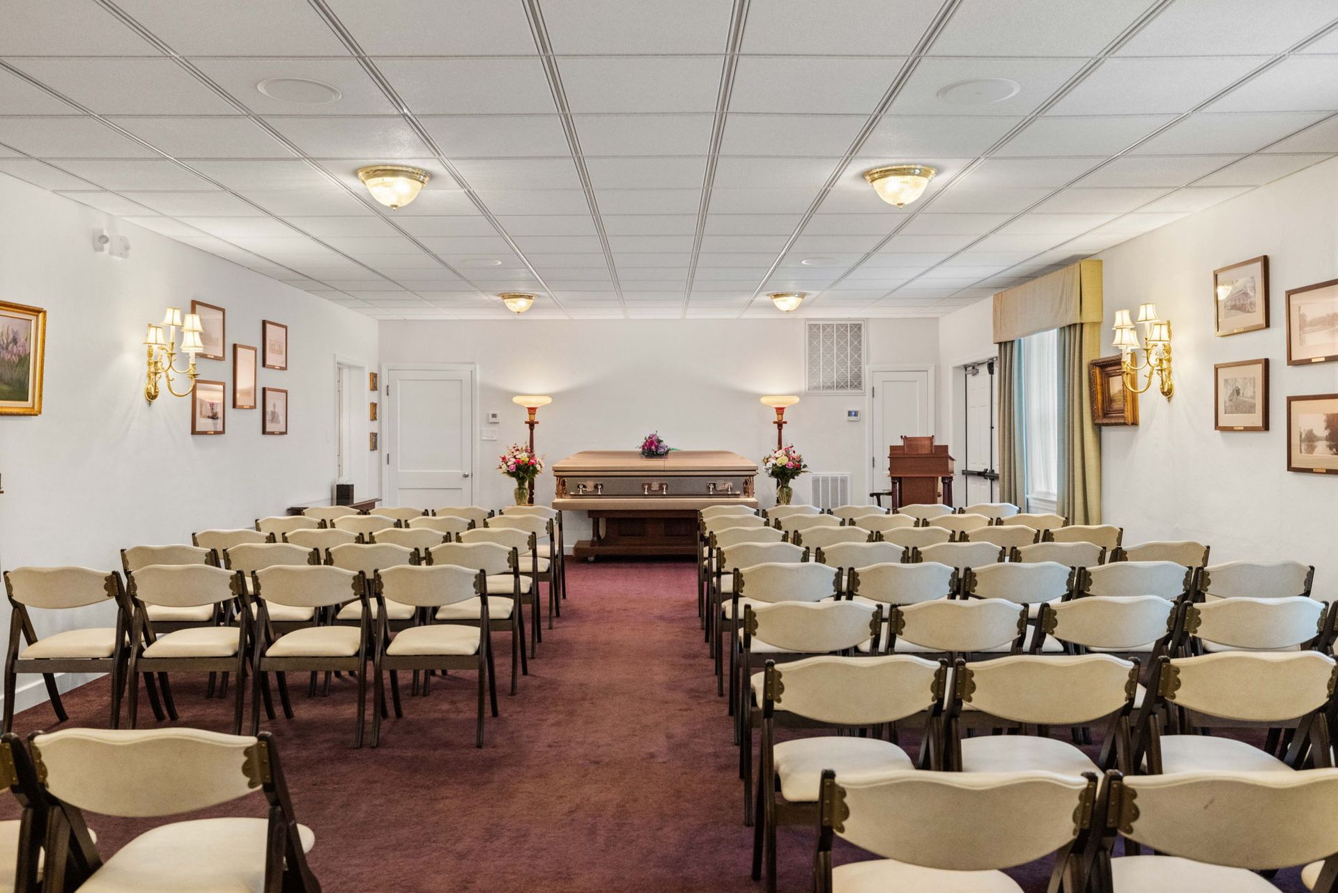 Funeral room with rows of beige chairs facing a front table and podium, lit by wall lamps