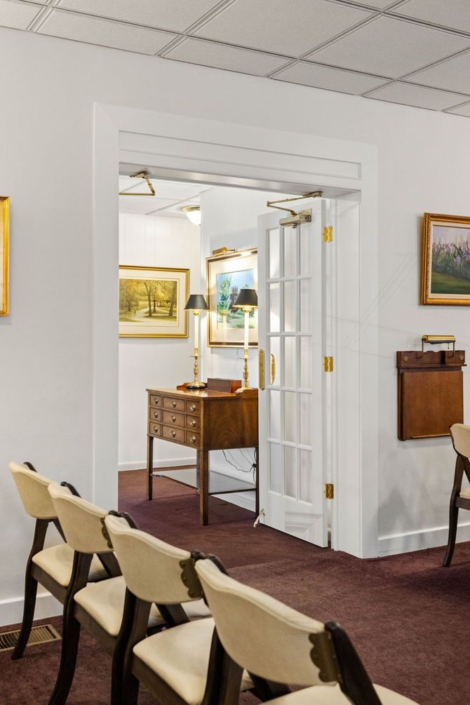 Empty gallery room with framed paintings, chairs, and open white double doors leading to another room