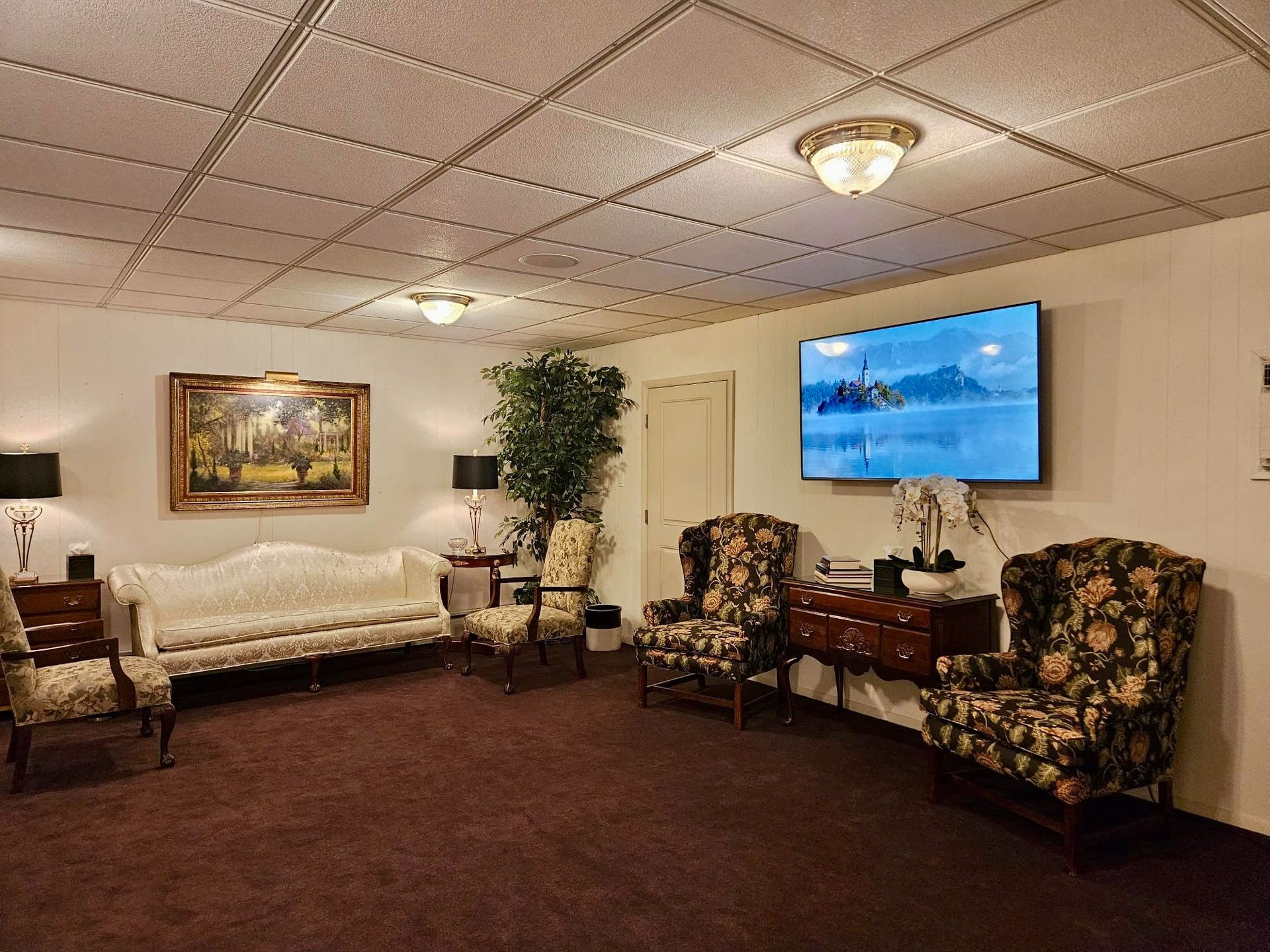 Quiet waiting room with white couch, floral chairs, potted plant, and wall-mounted TV on beige walls