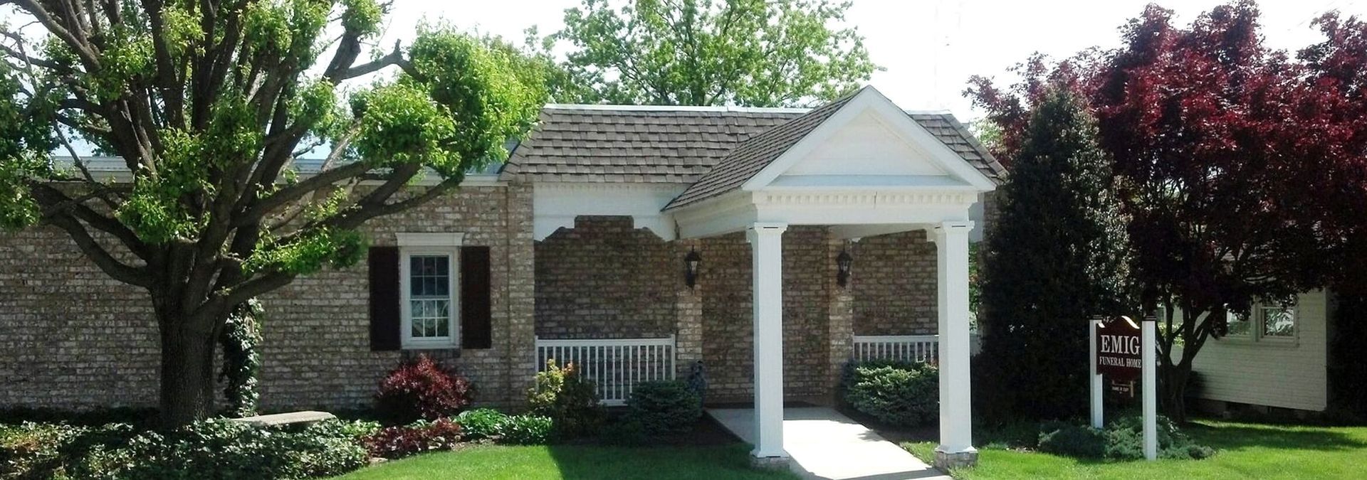 Small brick house with white porch and columns, surrounded by trees and shrubs.