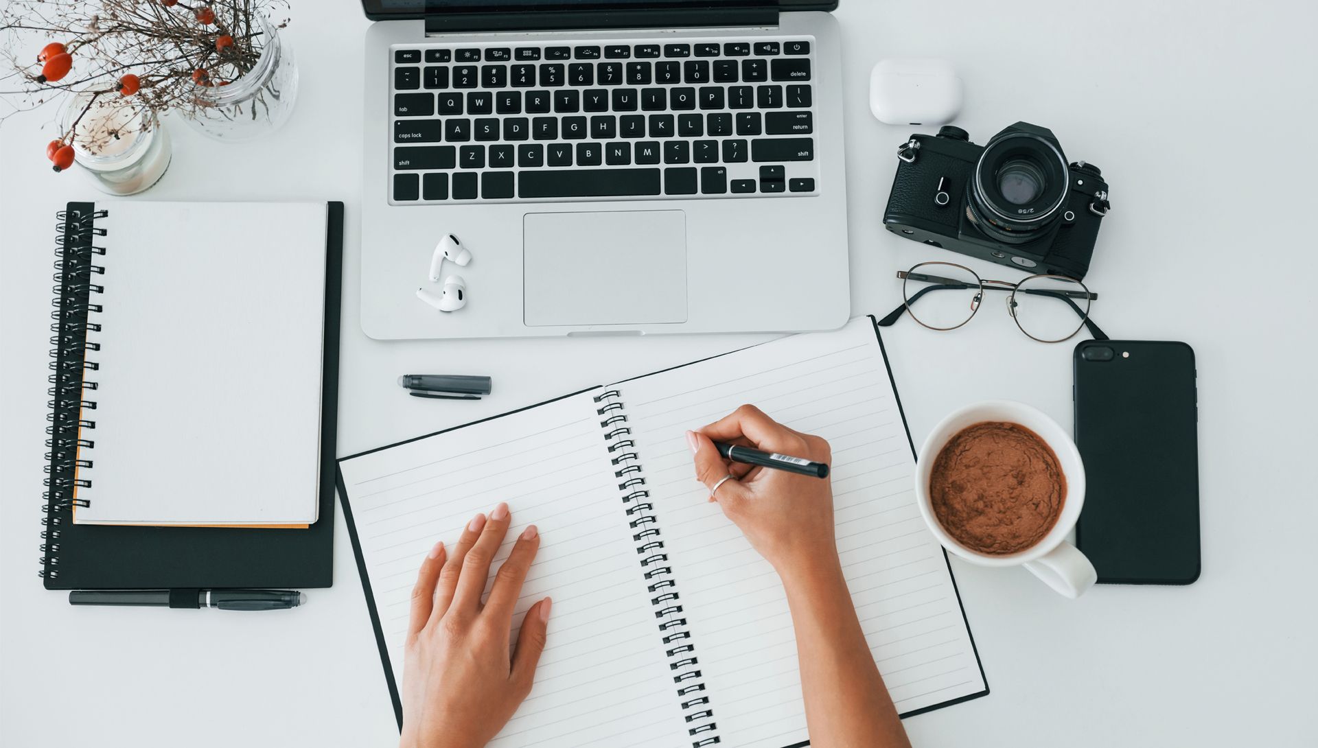 Person writing blog ideas in a notebook on a desk with a laptop, camera, coffee, and phone.