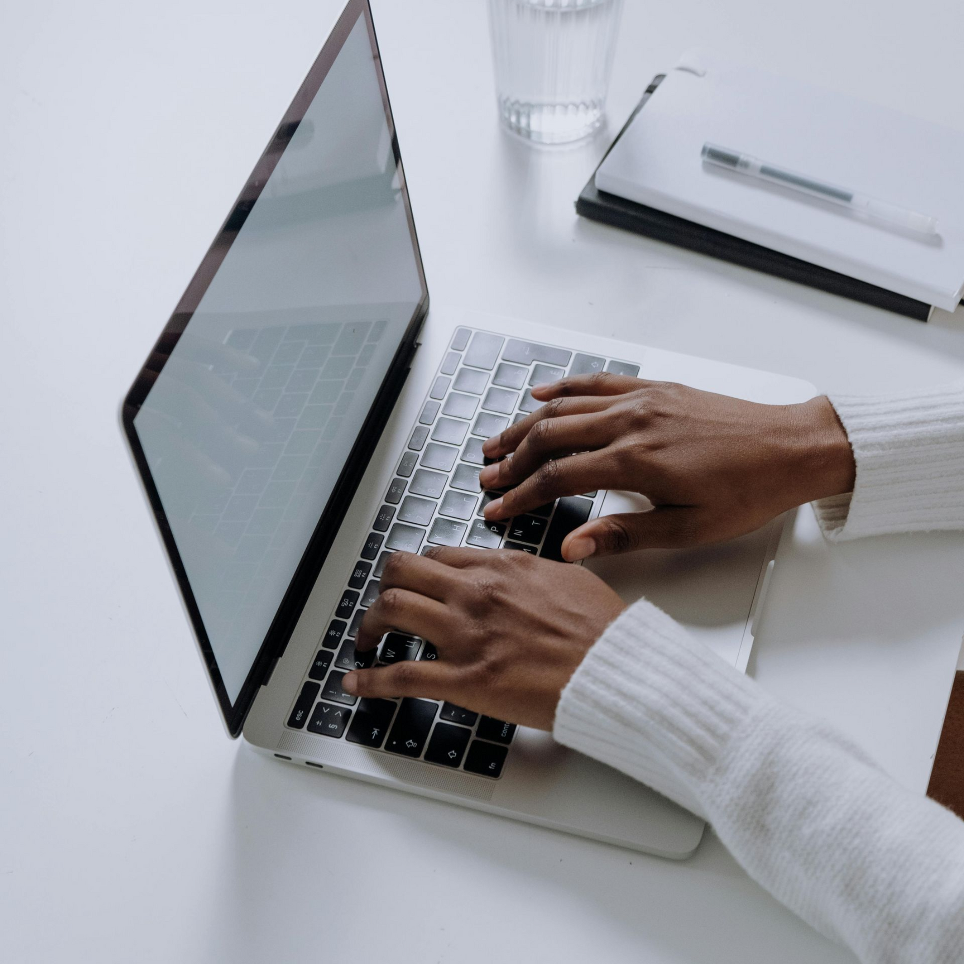 Hands typing on a laptop keyboard; a notebook and glass of water sit nearby on a white desk.
