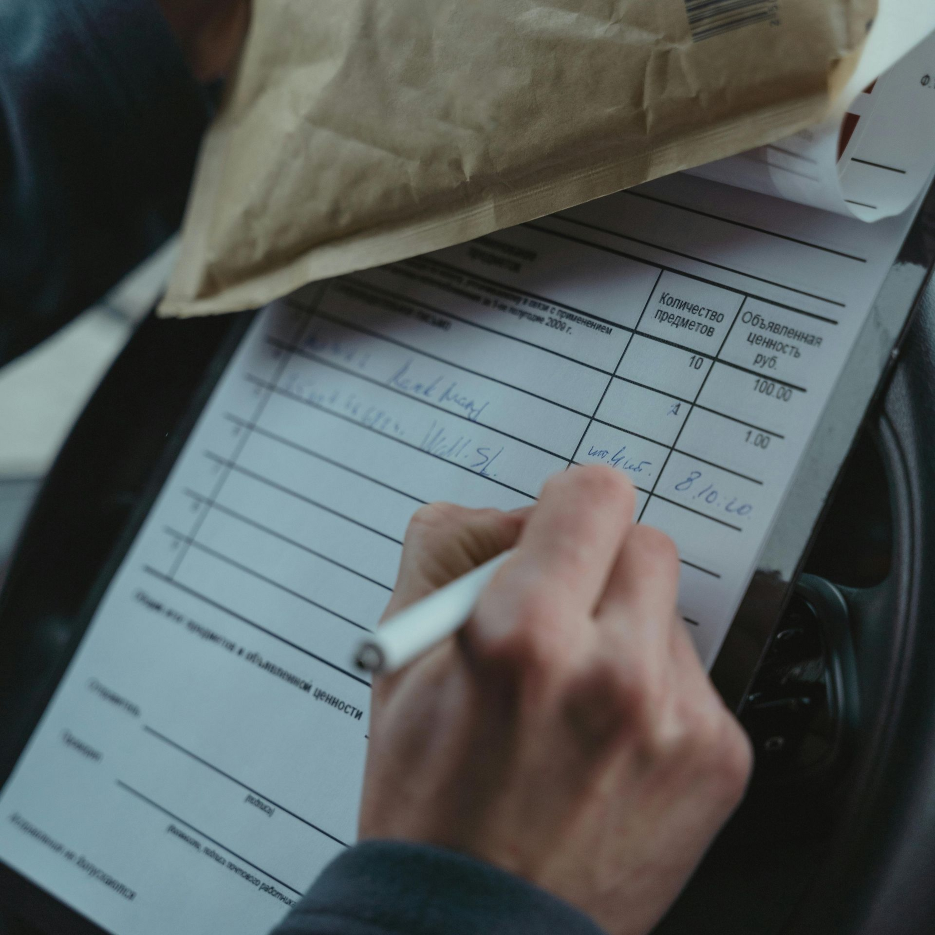 Person's hand writing on a form inside a vehicle; a brown paper bag is on top.