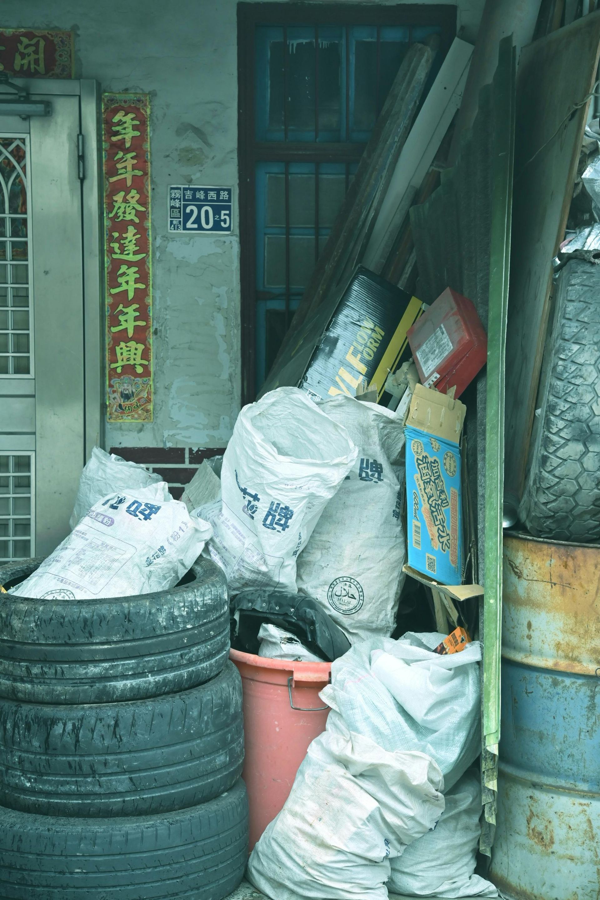 A cluttered alleyway next to a building.  White bags, tires, and debris are piled against a wall with a window and a doorway.