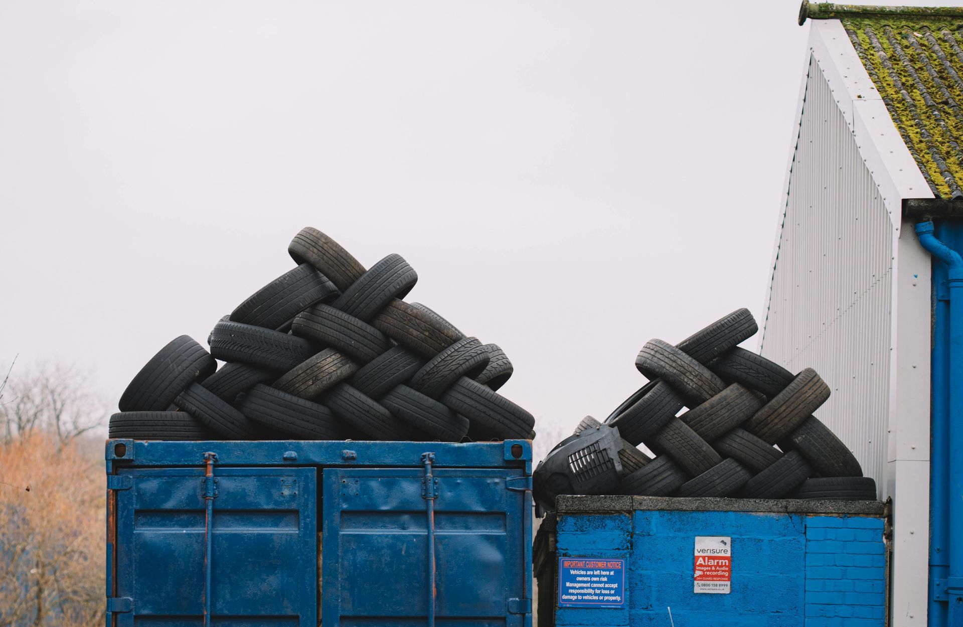 Blue containers overflowing with a pile of used tires, stacked on top. A white building is to the right.