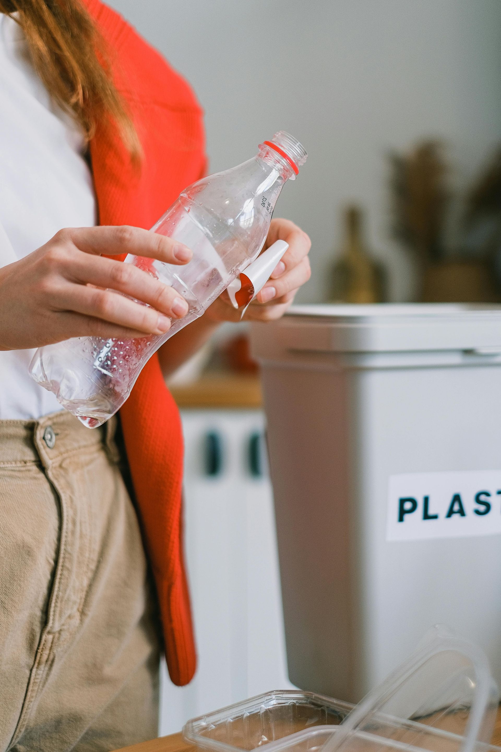 Woman putting a plastic bottle into a recycling bin. She wears a red jacket and tan pants.